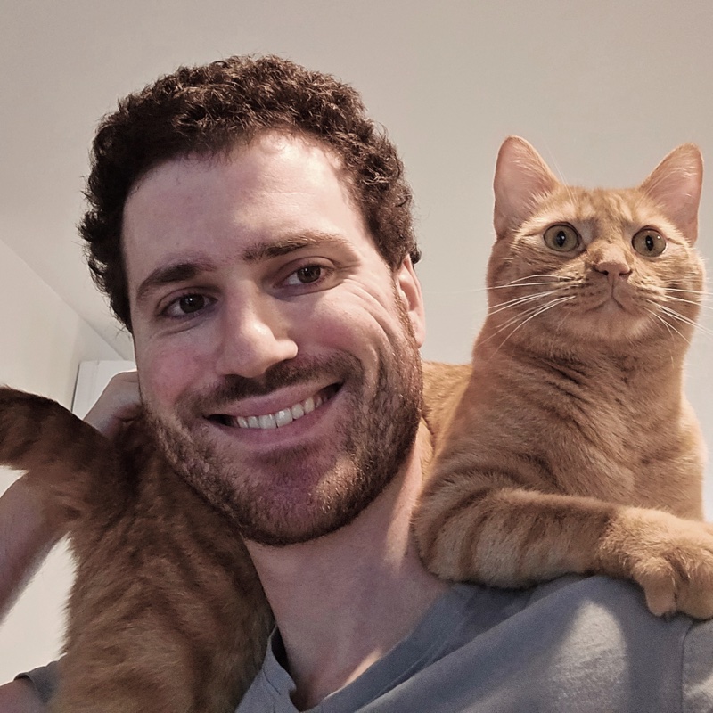 Graham Fluster smiling with his orange cat Pabu perched on his shoulder